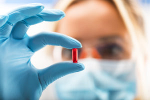 Young attractive female scientist with protective eyeglasses and mask holding a red transparent pill with fingers in gloves in the pharmaceutical research laboratory